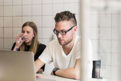 Young businessman working on laptop with female colleague having glass of water in new office