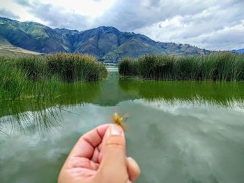 Cropped image of hand with reflection in lake against sky