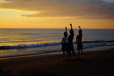 Silhouette friends with hand raised standing at beach against sky during sunset