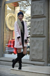 Beautiful woman in warm clothes standing against building on sidewalk