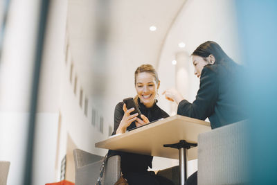 Low angle view of smiling businesswoman sharing smart phone with female coworker at table in cafeteria