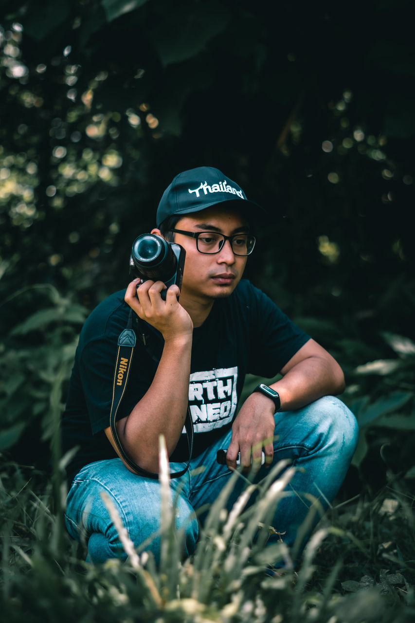 plant, blue, one person, forest, nature, adult, men, land, tree, glasses, green, outdoors, leisure activity, sitting, front view, clothing, person, lifestyles, casual clothing, fashion, cap, portrait, day, young adult, activity, grass