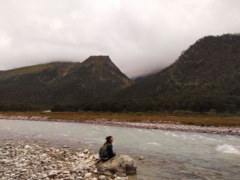 Woman sitting on mountain by lake against sky