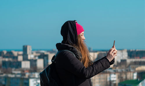 Young woman in pink hat with smartphone calling friends walking on city background millennial girl 