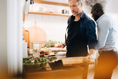 Mature man working at counter while standing with friend in kitchen