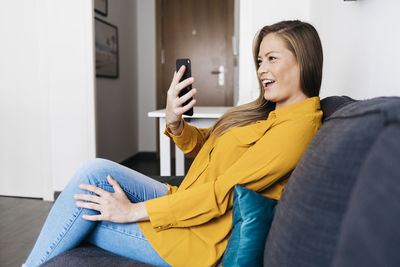 Happy young woman taking selfie with mobile phone while sitting on sofa at home