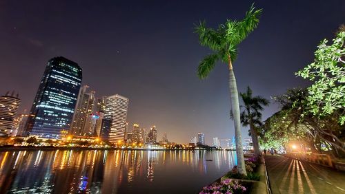 Illuminated buildings by palm trees against sky at night