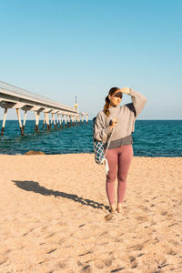 Full body of young barefoot female in activewear with rolled yoga mat looking at distance while standing on sandy beach near sea