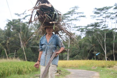 Portrait of woman holding wood on head while walking on footpath against sky