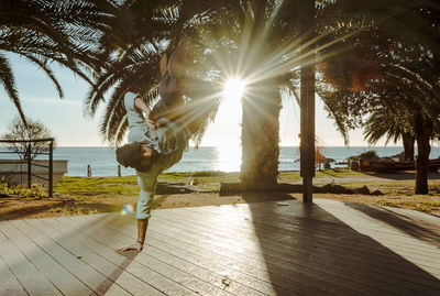 Contemporary male dancer in stylish casual clothing standing on hand with crossed legs above head at outdoors terrace of house with tropical palms seashore and sun beams on background