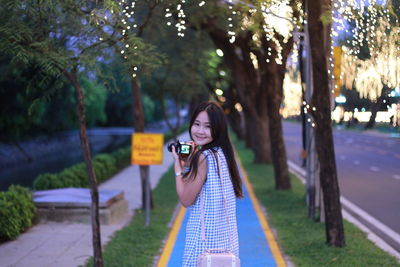 Portrait of young woman holding camera and standing at footpath