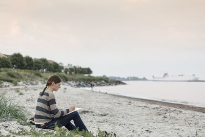 Woman writing on book while sitting at beach against sky