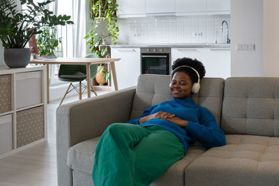 Happy relaxed african american woman in headphones closing eyes enjoying music sits on sofa