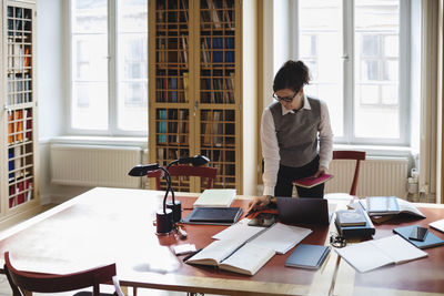 Female professional holding book while standing at table in library