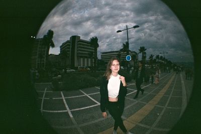 Portrait of beautiful woman standing on footpath against cloudy sky