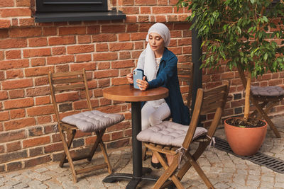 Young woman sitting on chair against brick wall