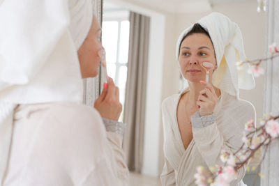 Portrait woman wrapped in white towel and in bathrobe looks at reflection