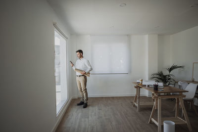 Architect holding rolled up blueprint using smart phone standing by window in office