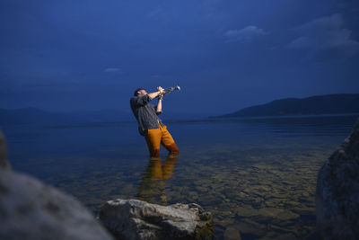 Man playing trumpet while standing in sea against sky at dusk