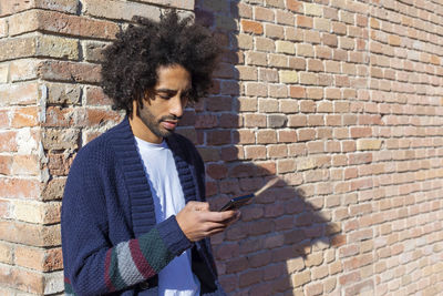 Young man using phone while standing against brick wall