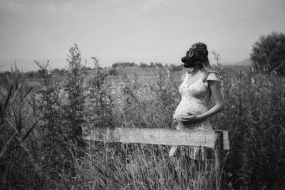 Pregnant woman wearing gas mask while standing by plants against sky