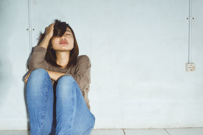 Sad young woman with eyes closed sitting against wall at home
