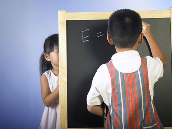 Siblings writing on blackboard while standing against blue background