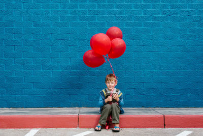 Boy with red balloons sitting on sidewalk against blue wall