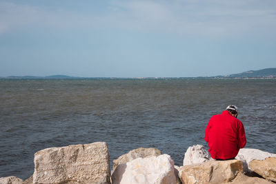Rear view of man crouching on rock by sea against sky