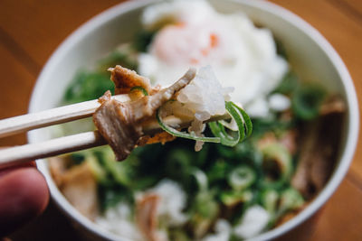 Cropped hand of person holding food in chopsticks against bowl