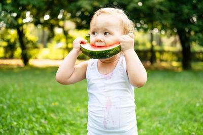 Funny smiling kid boy in white bodysuit eating watermelon at green lawn