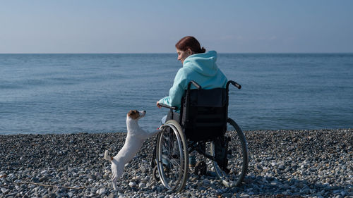 Caucasian woman in a hoodie in a wheelchair with a dog on the seashore.