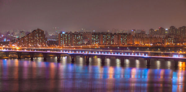 Illuminated bridge over river by buildings against sky at night