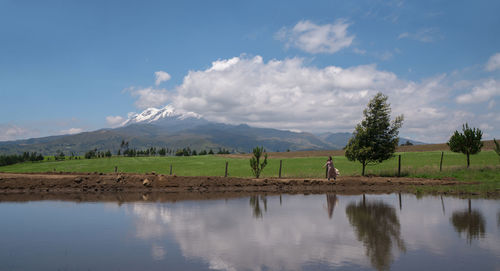 Woman walks with hat along a dirt road reflecting in lagoon with mountain during a sunny morning