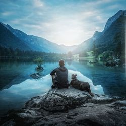 Man with dog sitting on rock at lakeshore against mountains