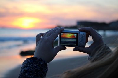 Cropped image of woman hand photographing sea against sky during sunset
