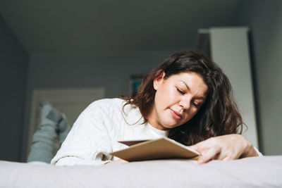 Young adult brunette woman plus size body positive in sweater writes notes in planner on bed at home