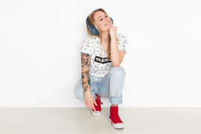 Smiling woman listening to music while crouching against wall