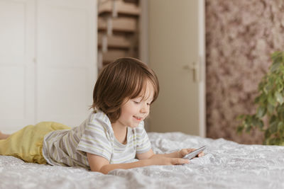 A cute boy, toddler, lies on the bed in the bedroom and plays with the phone, smartphone. 