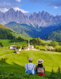 Rear view of woman sitting on grassy field by mountains against sky