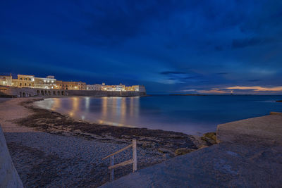 Illuminated city by sea against blue sky at dusk