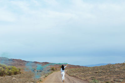 Rear view of woman holding distress flare while running on dirt road against cloudy sky