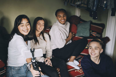 Smiling teenage girl taking selfie with friends on monopod while enjoying at home