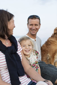 Cheerful girl sitting amidst father and mother with dog against sky