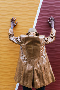 Back view of anonymous male in stylish leather coat and hat leaning hands on colorful wall while standing on street