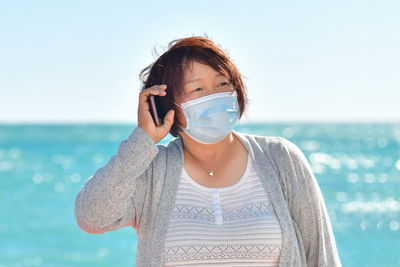 Mature woman wearing mask talking on phone while standing against sea
