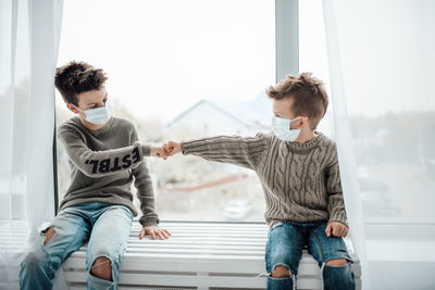 Brothers wearing mask doing fist bump sitting by window at home