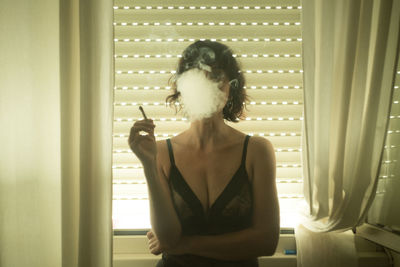 Woman smoking cigarette against window at home