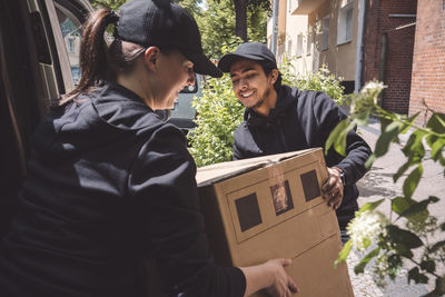 Smiling delivery colleagues holding cardboard box near truck