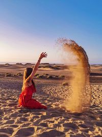 Full length of woman throwing sand mid air while sitting on sand against sky